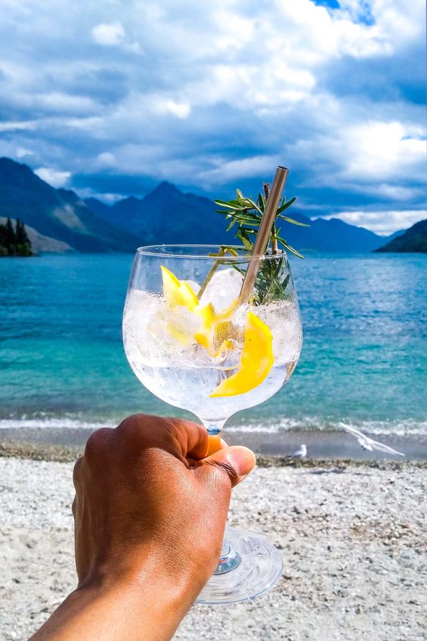A glass of gin and tonic with a sprig of rosemary in front of an aqua blue lake and mountains